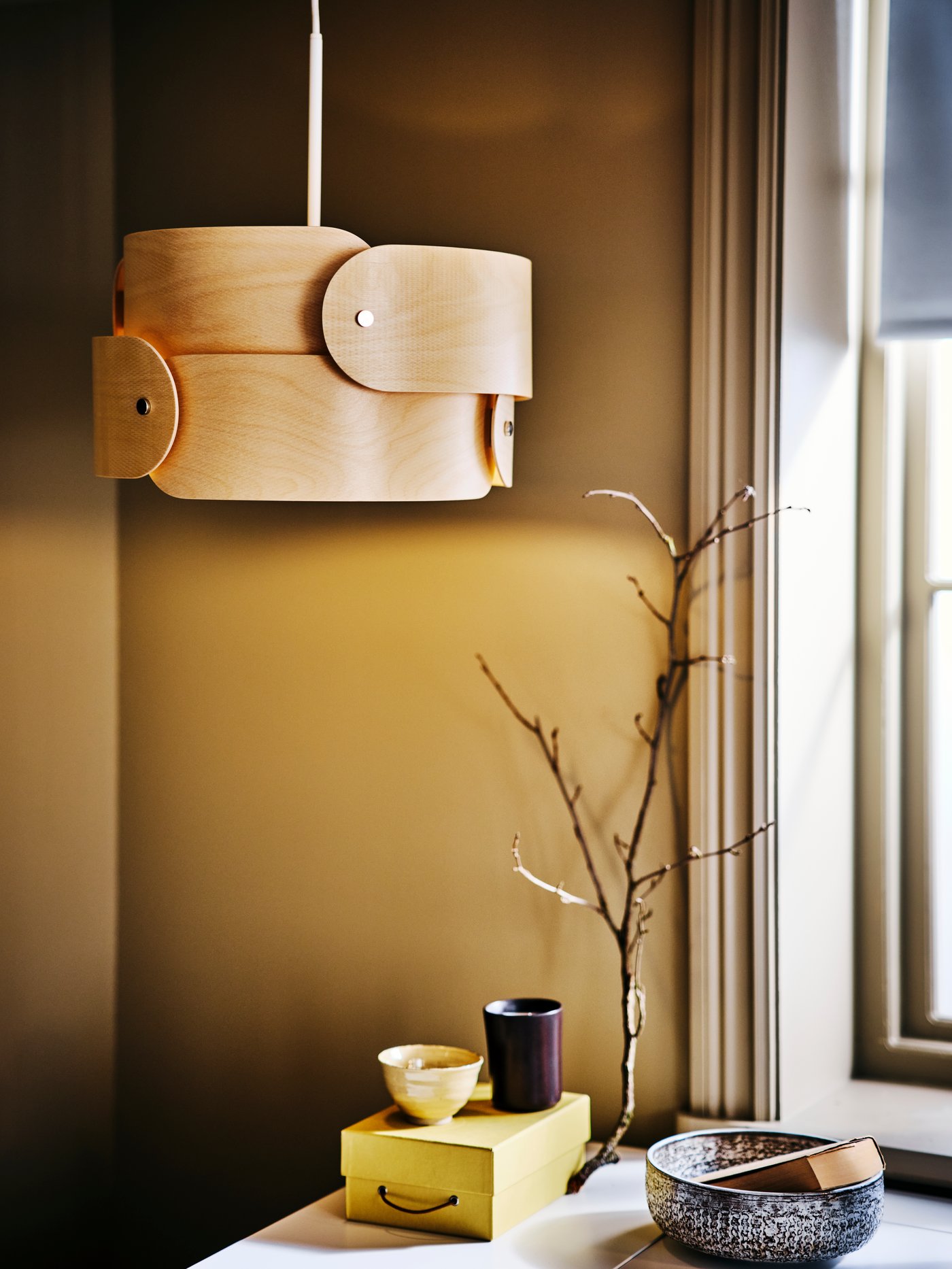 A lit SÖDÅKRA pendant lamp in the corner of a room, with ornamental bowls, a box and a branch atop a surface below.
