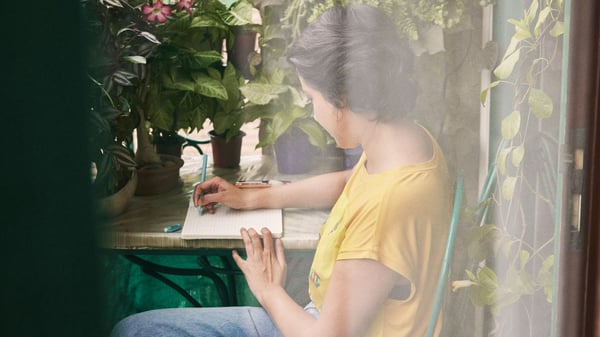 Mulher com t-shirt amarela sentada à mesa no seu jardim a escrever num caderno.