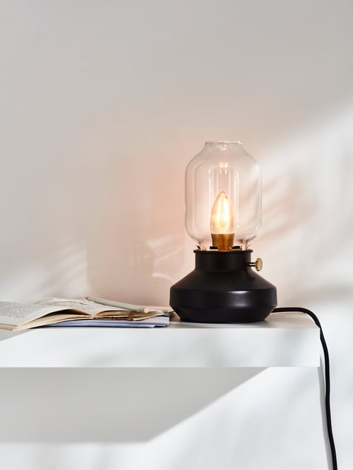 An anthracite table lamp with a glass shade and a lit, visible bulb sits on a white shelf by open books and a pencil.