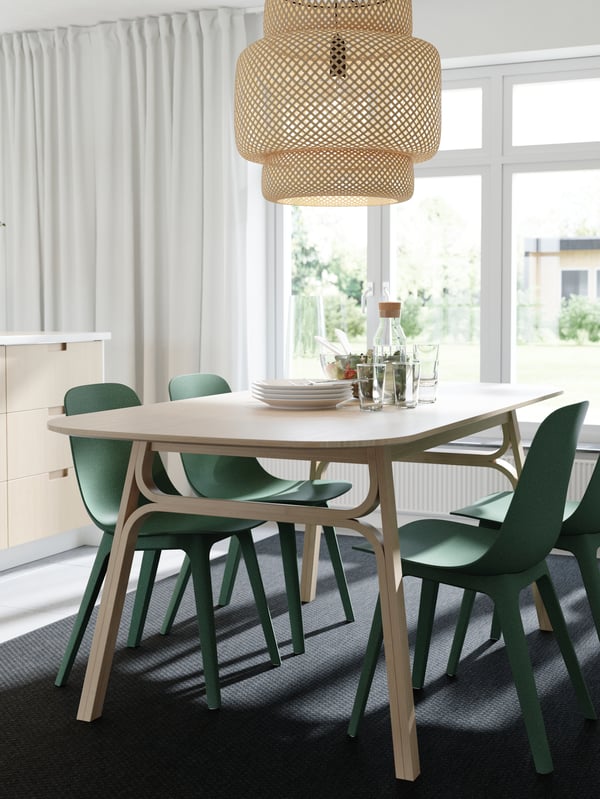 A dining table, a pendant lamp and kitchen drawer fronts in bamboo, green chairs, a dark grey rug and white curtains.
