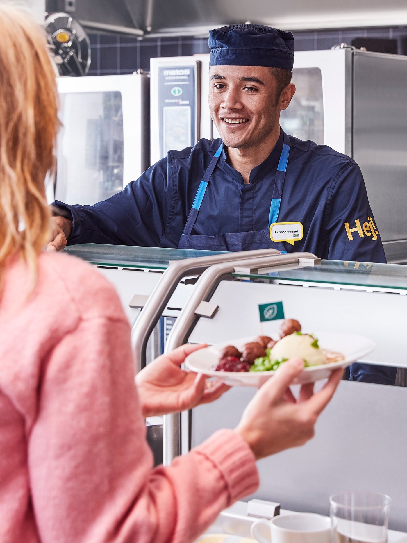 A customer wearing a pink jumper collects her meal from a smiling Swedish bistro co-worker wearing a dark blue uniform.
