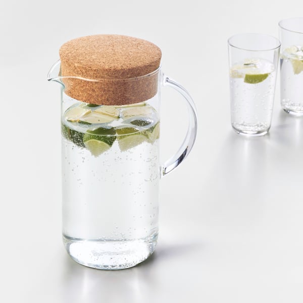 Pieces of lime float in water inside a glass IKEA 365+ jug with cork lid and a tall glass behind it, both on a white surface.