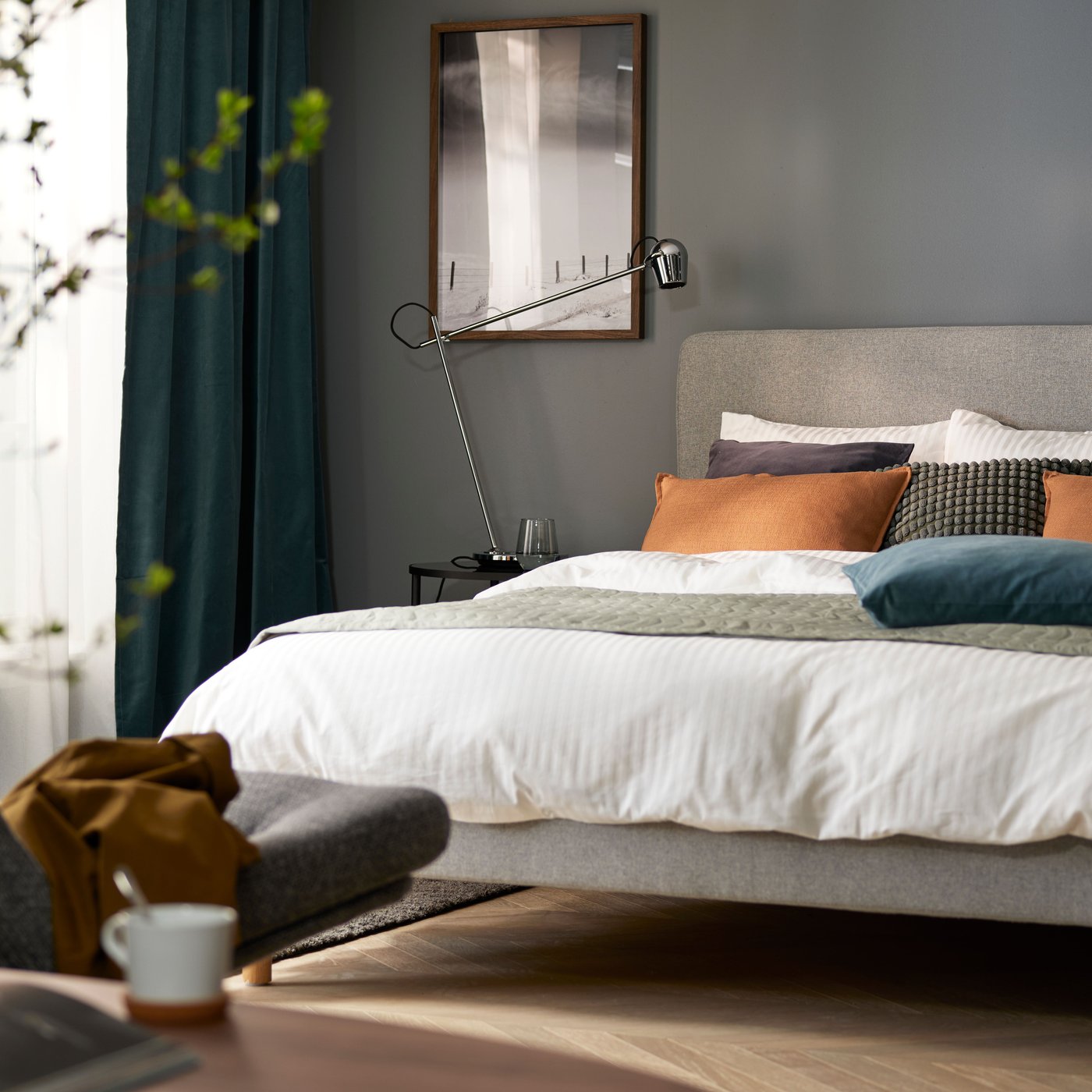 A bedroom with dark grey walls and a bed with white-grey-orange bedding, an armchair in foreground and windows in back.