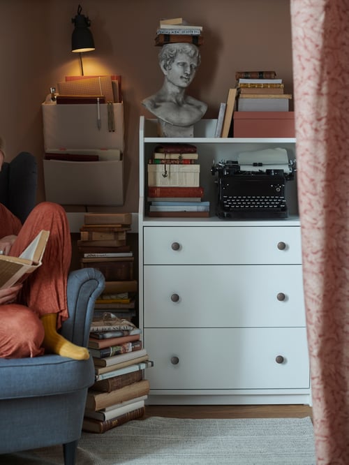 A white bookcase with drawers and shelves propped with old books, a typewriter and sculpted bust placed by a reading corner.
