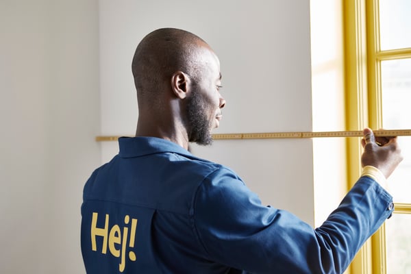 Un collègue d'IKEA portant une veste bleue mesure un mur à côté de la fenêtre.