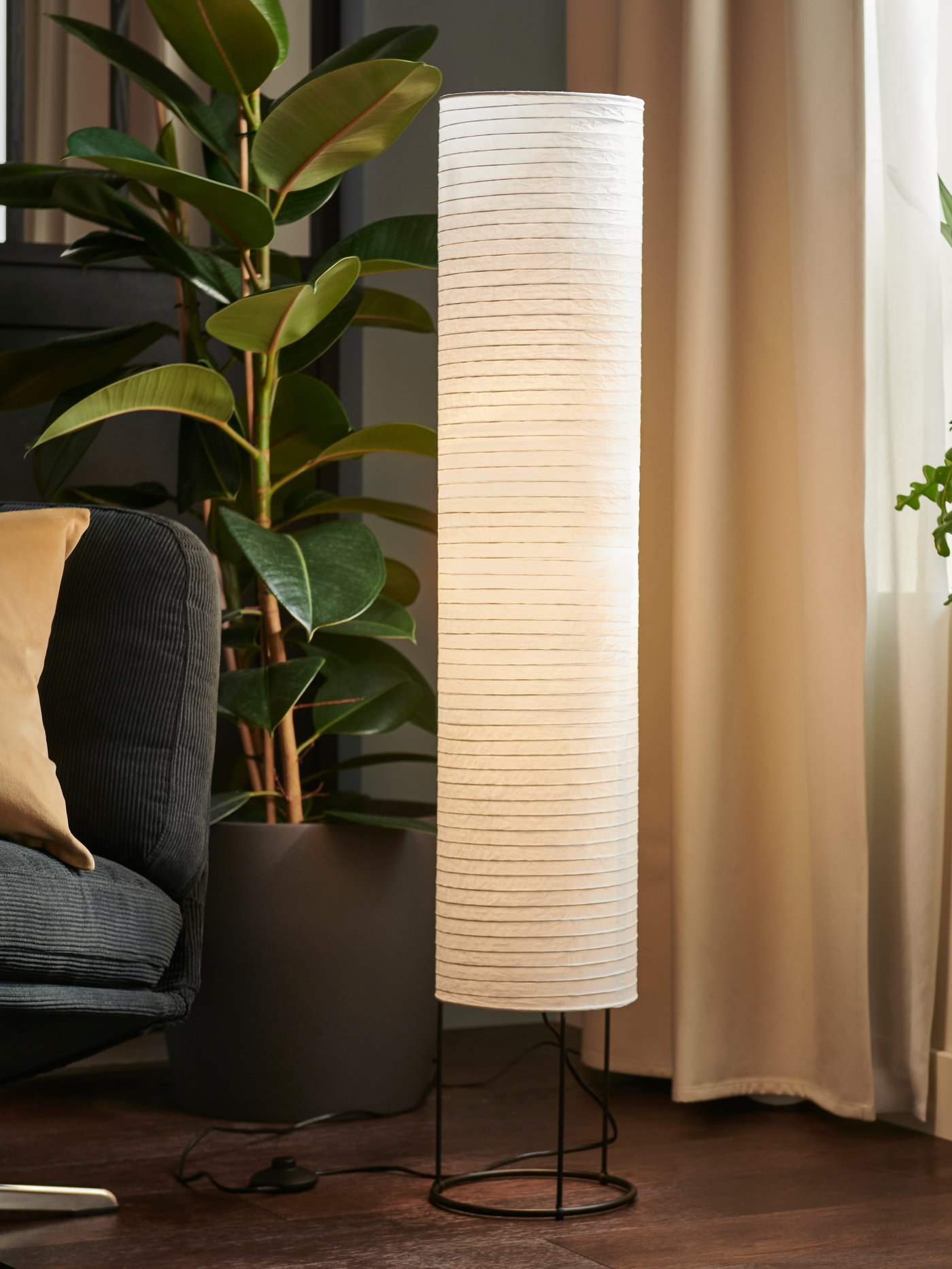 A corner of a living room furnished with a DYVLINGE swivel armchair, a large leafy plant, and a lit STRANDAD floor lamp.
