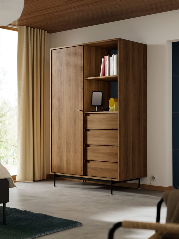 Geöffneter RÅDMANSÖHolzkleiderschrank mit Regalböden, auf denen Bücher und weitere Gegenstände liegen; darunter vier Schubladen. Im Raum sind helle Vorhänge, ein Fenster und links ein Teil eines Bettes zu sehen.