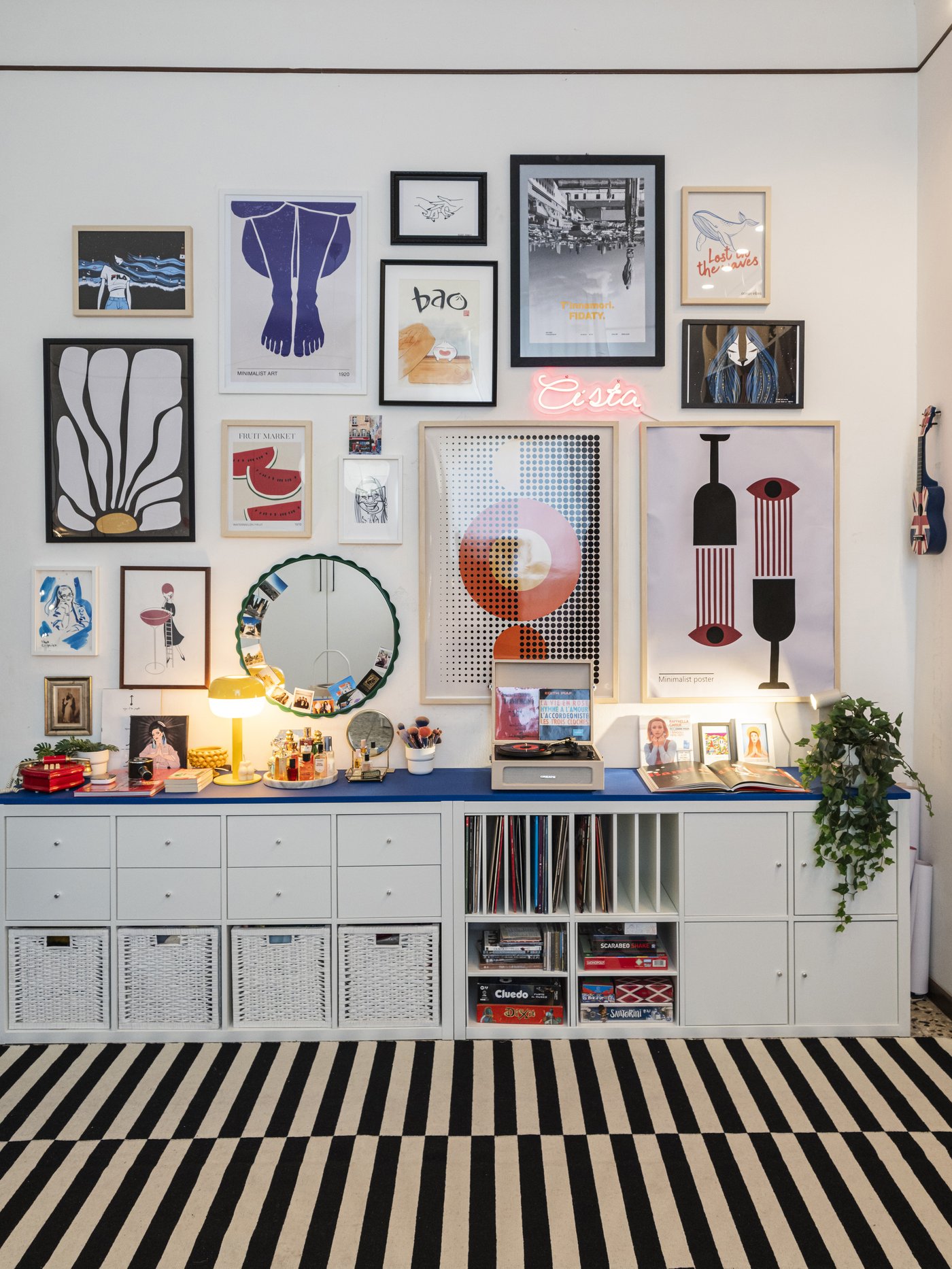 Parete decorata con una galleria di quadri colorati sopra moduli contenitore KALLAX bianchi, con piano d’appoggio blu e oggetti decorativi, su tappeto a righe bianche e nere.
