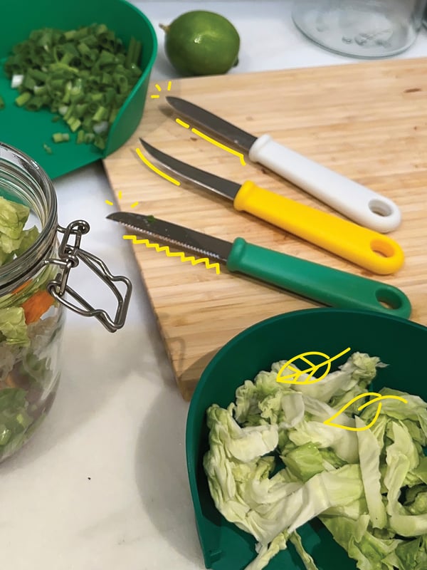 Three paring knives on a chopping board, with chopped vegetables placed in measuring bowls around it.