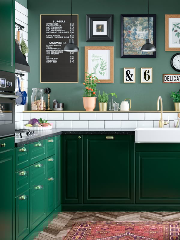 Angolo di una cucina sui toni del verde e piastrelle bianche. Sulla parete, cornici e stampe.