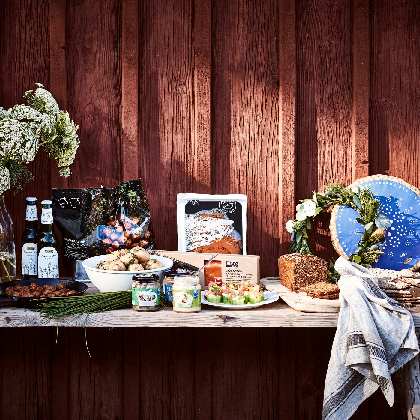 Reich gedeckter Holztisch mit Brot, Käse, Getränken, Blumen und verpackten Lebensmitteln vor einer roten Holzfassade.
