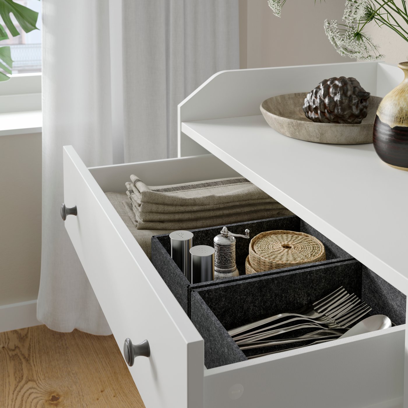 The open drawer of a HAUGA chest of 6 drawers in white contains folded cloth napkins, coasters and cutlery.