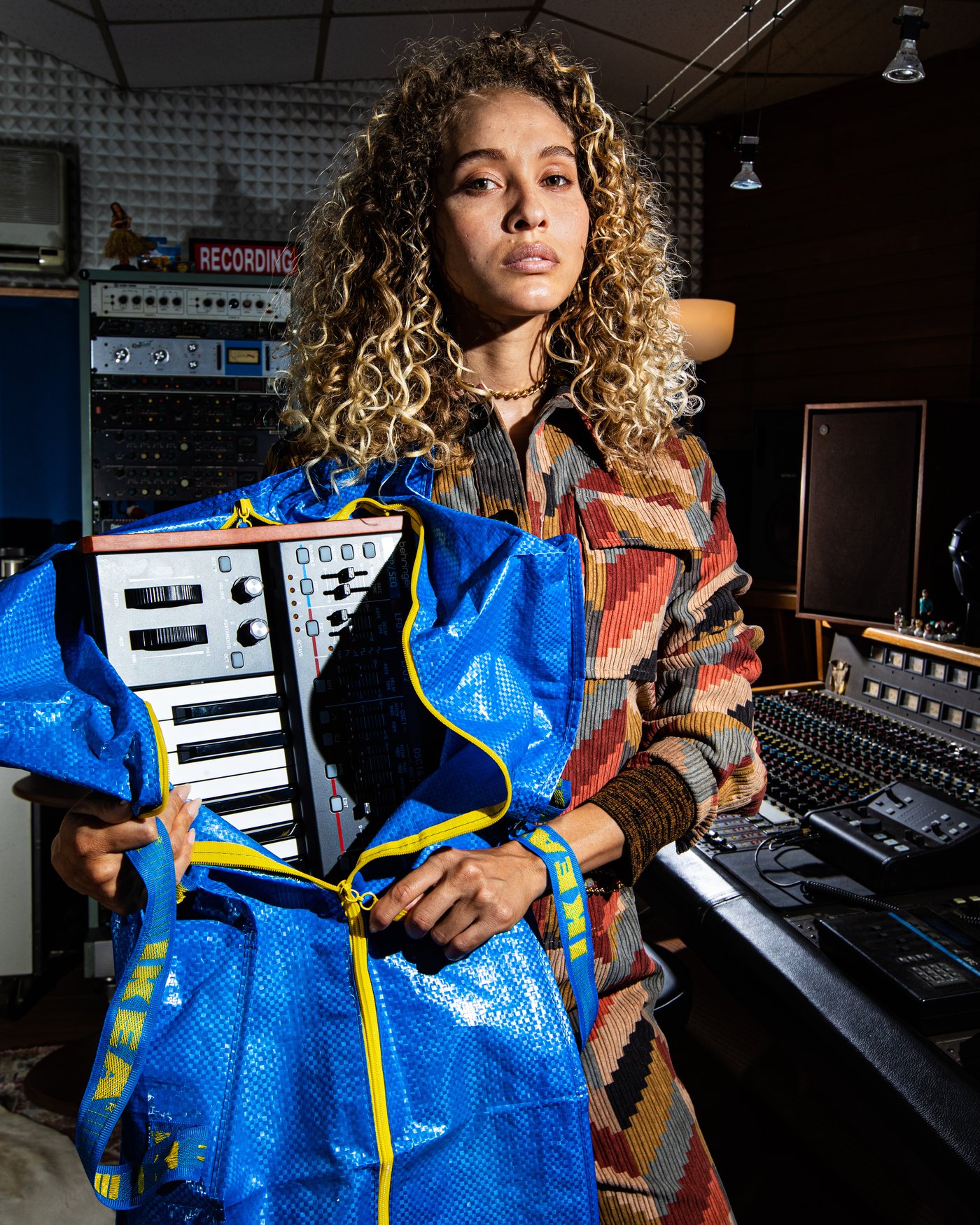 Donna riccia con camicia fantasia regge una borsa blu FRAKTA IKEA da cui fuoriesce un sintetizzatore a tastiera in uno studio di registrazione.