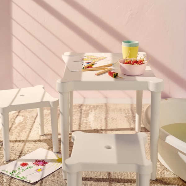 Un tavolino con alcuni sgabelli intorno. Sopra piatti e bicchieri KALAS con alimenti e spiedini di anguria. Ci sono anche fogli e pastelli per colorare.