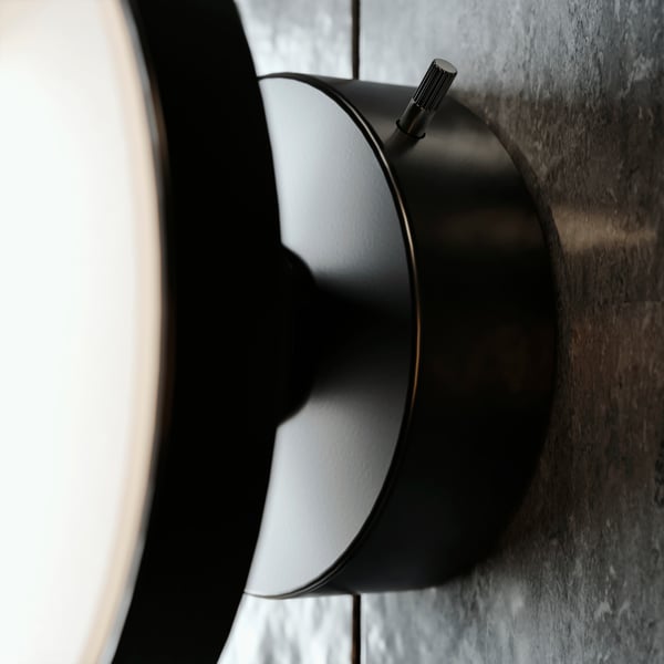 A close-up of a matte black KABOMBA LED lamp mounted on a dark grey wall with a button that can be used to dim the light.