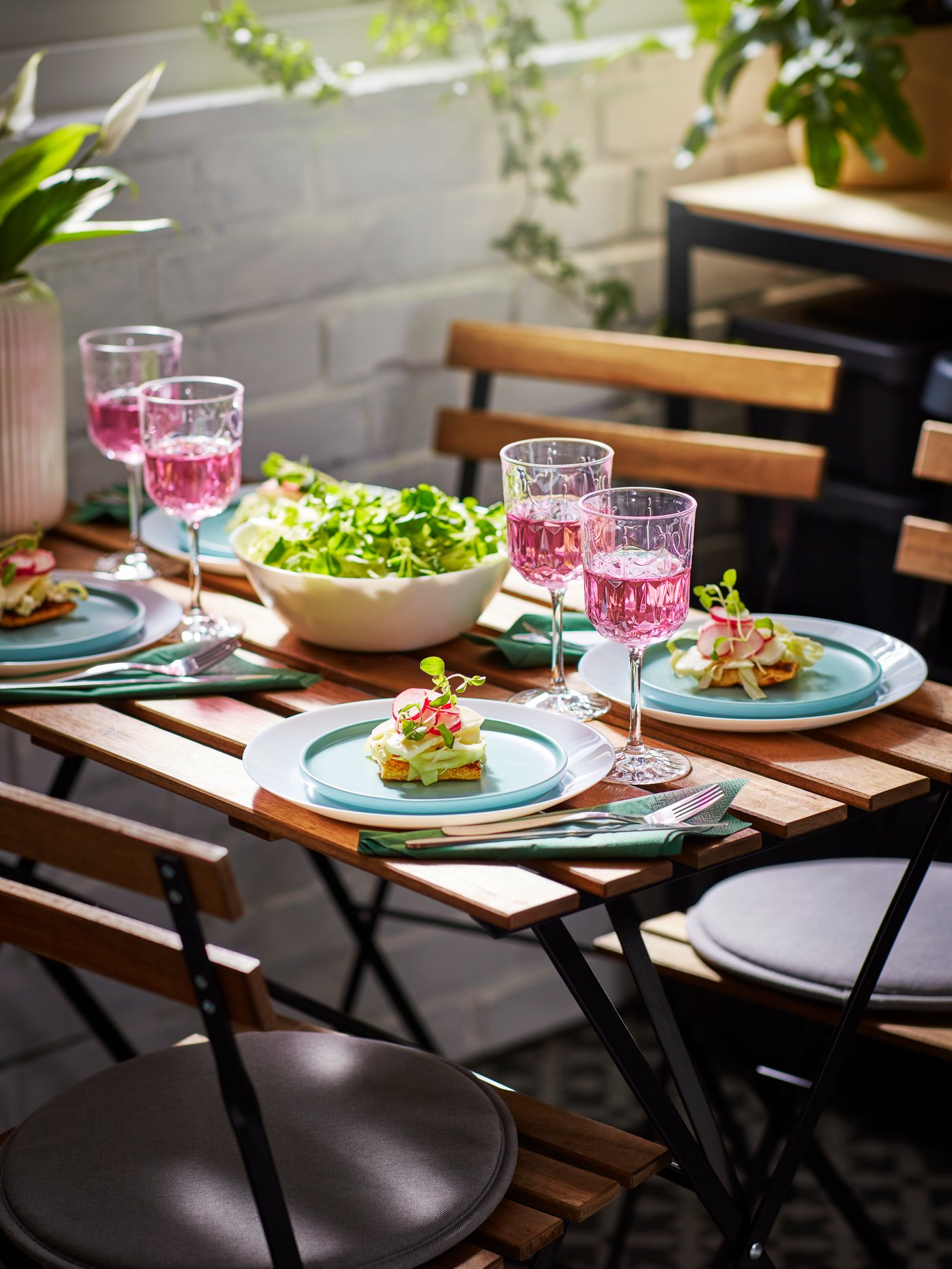 Mesa de jardín para comer; copas de vino, ensalada en un cuenco OFTAST blanco y entrantes en platos KEJSERLIG de color turquesa.