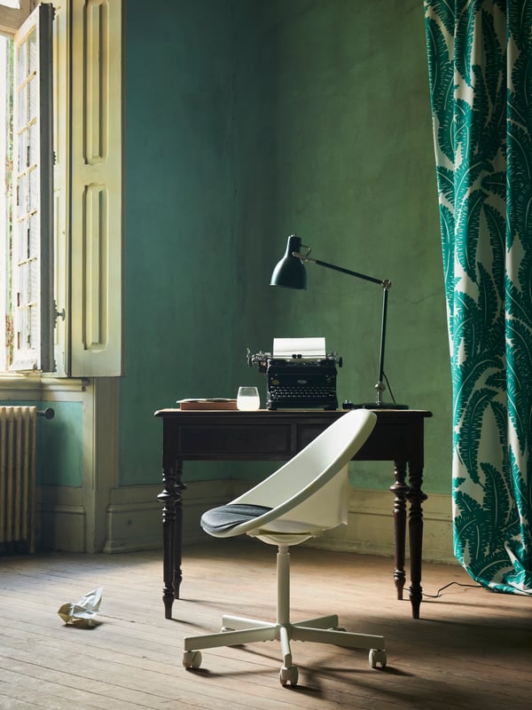 The white LOBERGET/BLYSKÄR swivel chair with a rounded design placed by an old desk near an open window in a green room.