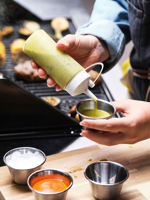 Someone is squeezing green sauce from a white/transparent GRILLTIDER squeeze bottle into a sauce cup.