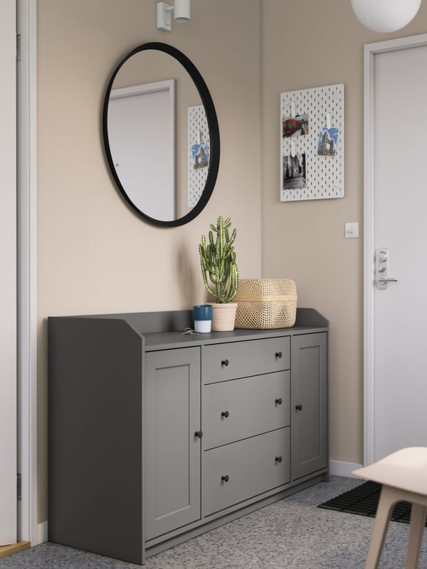 A grey HAUGA sideboard by a front door in a hallway, with a round mirror above it and a plant and a box on top.