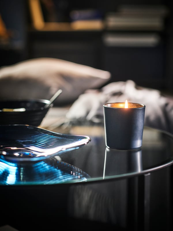 The top of a round VITTSJÖ coffee table with a burning FRUKTSKOG scented candle and a blue HÄCKPOPPEL decorative bowl on top.