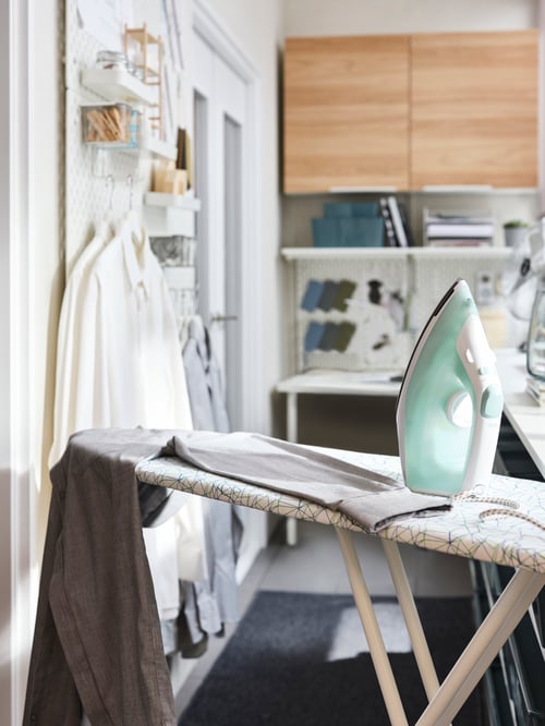 A modern laundry room that features an ironing board, a table top, a wall cabinet with two shelves, and a pegboard. 