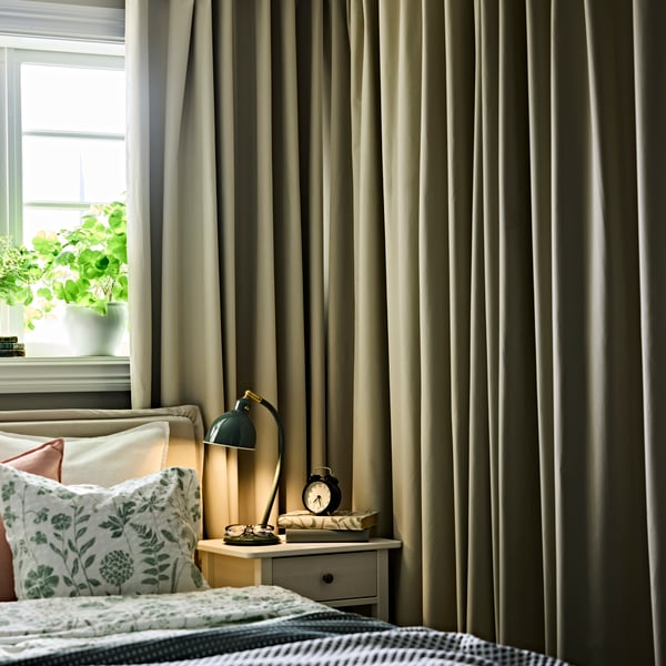 Chambre avec rideaux opaques BENGTA près d’un lit bien fait et d’une table de chevet accueillant une lampe et un réveil.