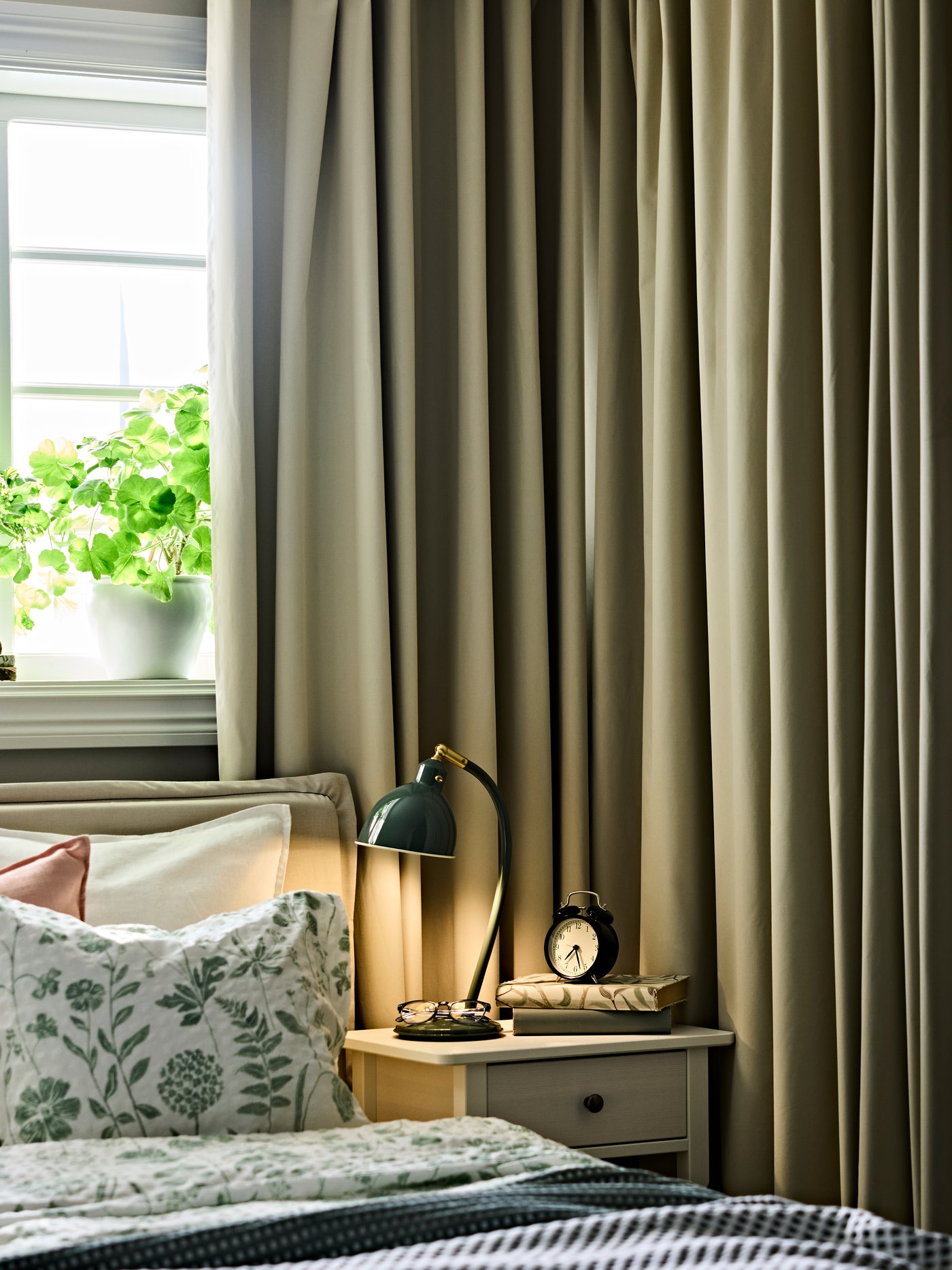 Ein Schlafzimmer mit BENGTA‑Verdunkelungsvorhängen neben einem ordentlich gemachten Bett sowie einem Nachttisch mit Lampe und Wecker.