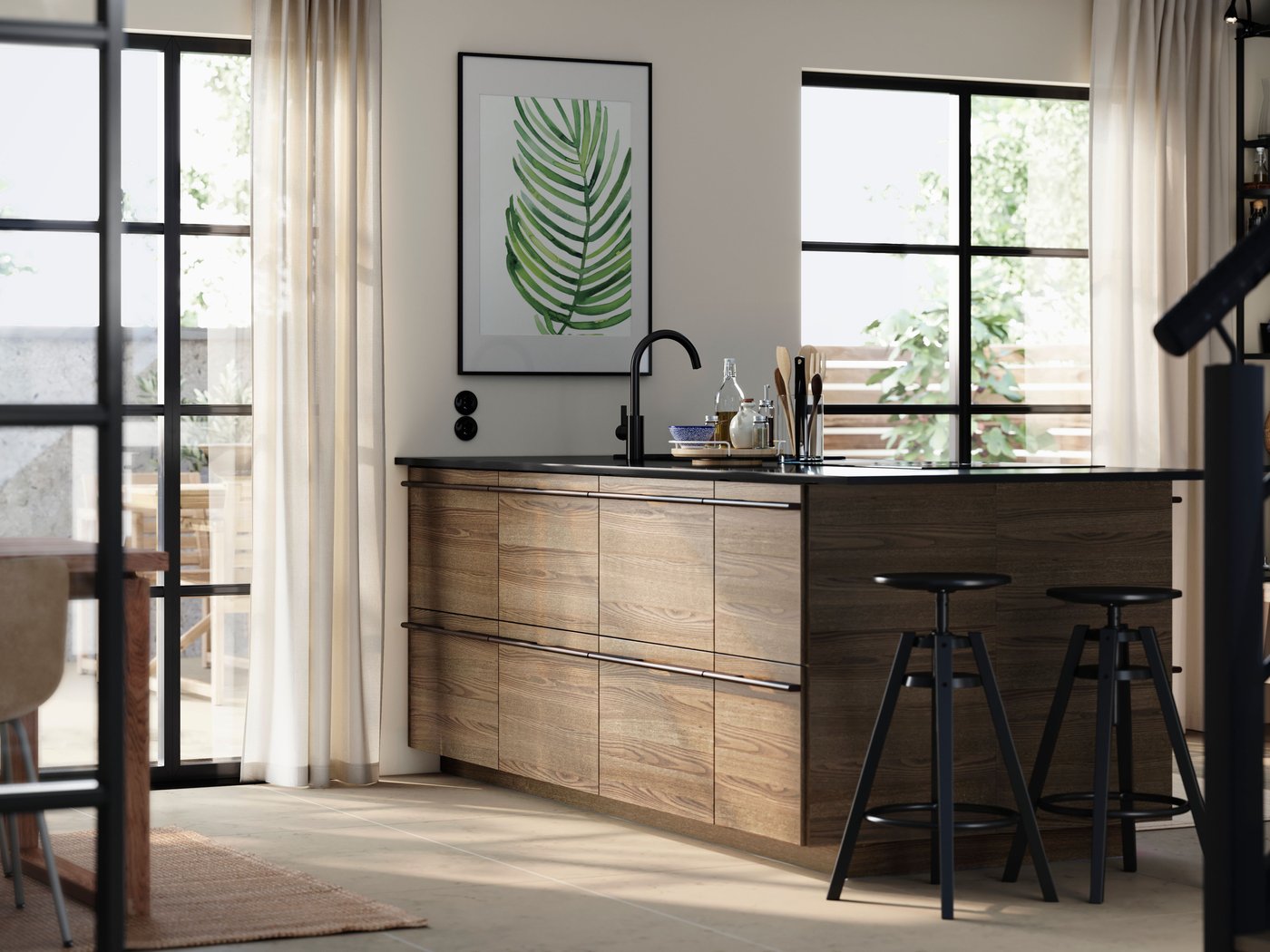 Un îlot de cuisine avec des façades en placage de frêne brun foncé de STENSTA, complété par des tabourets de bar et une grande plante imprimée sur le mur.