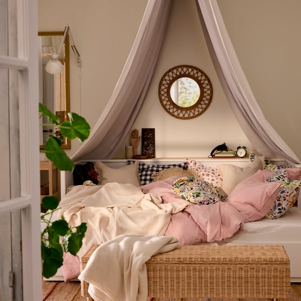 HEMNES day-bed with bedlinen in pink and white, TOLKNING rattan bench in foreground, a mirror and a textile canopy above bed.