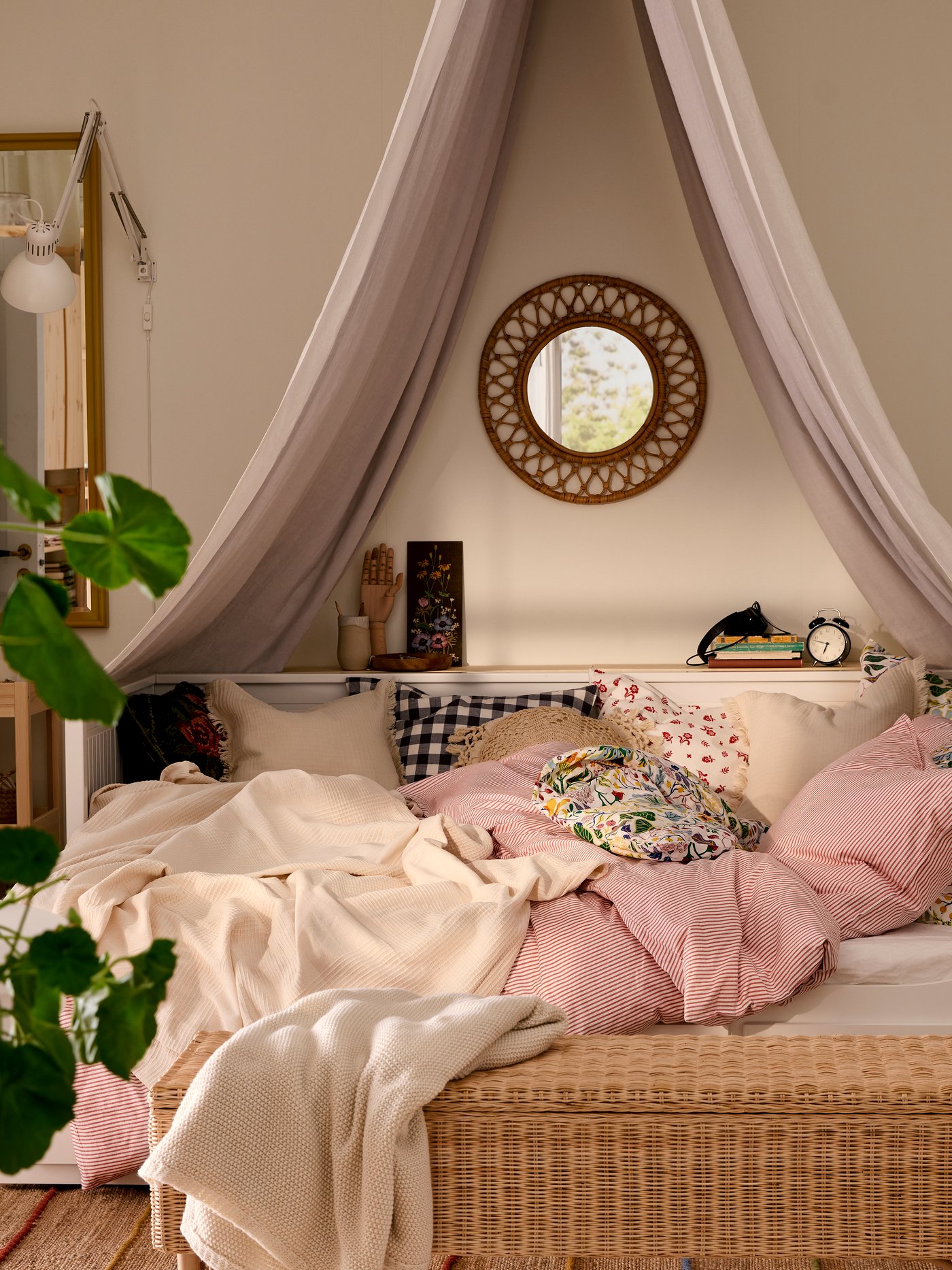 HEMNES dagbädd med sängkläder i rosa och vitt, TOLKNING bänk i rotting i förgrunden, en spegel och ett textilt sänghimmel ovanför sängen.
