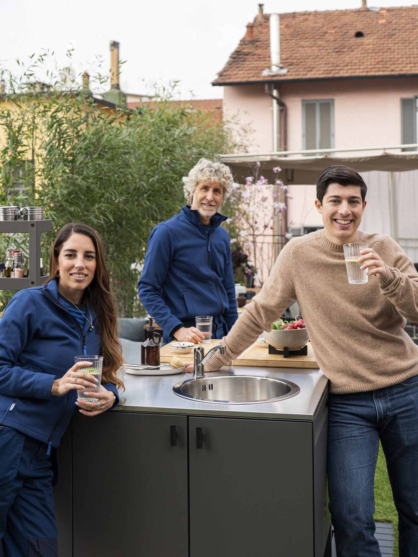 Marcello Ascani e due Interio designer IKEA sorridenti in un terrazzo, appoggiate a una cucina da esterno, mentre bevono da bicchieri davanti a una casa. 