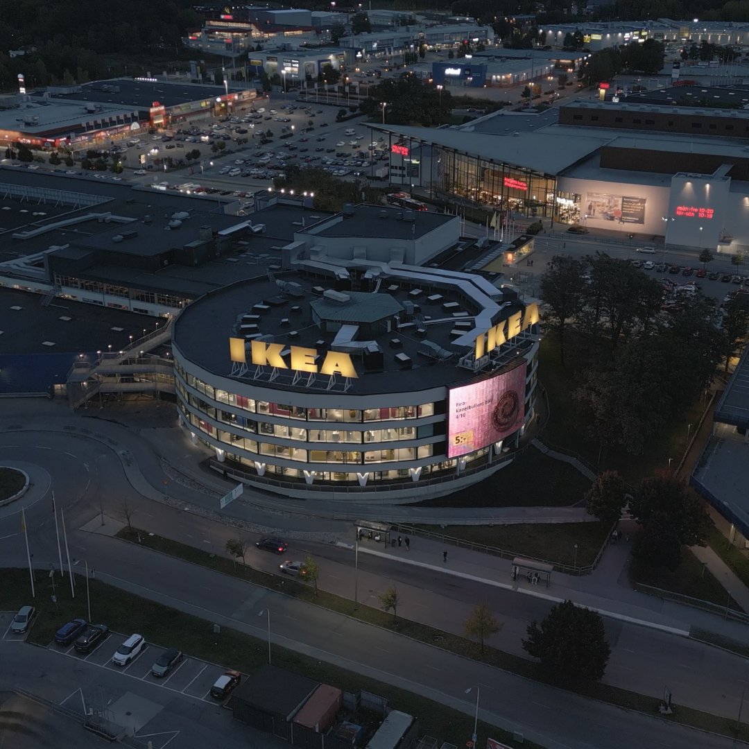 Flygbild i skymning över IKEA Kungens Kurva, ett stort cirkulärt varuhus med upplyst IKEA-logotyp på taket och fasaden. Byggnaden har flera våningar med stora fönster och en digital reklamskylt. Omkringliggande område visar parkeringsplatser, vägar och andra handelsbyggnader.