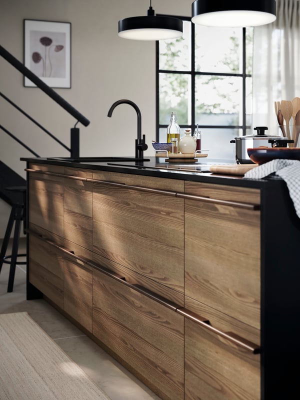 A kitchen with STENSTA dark brown ash veneer drawer fronts, black tap, black countertop, and cooking utensils placed on top.