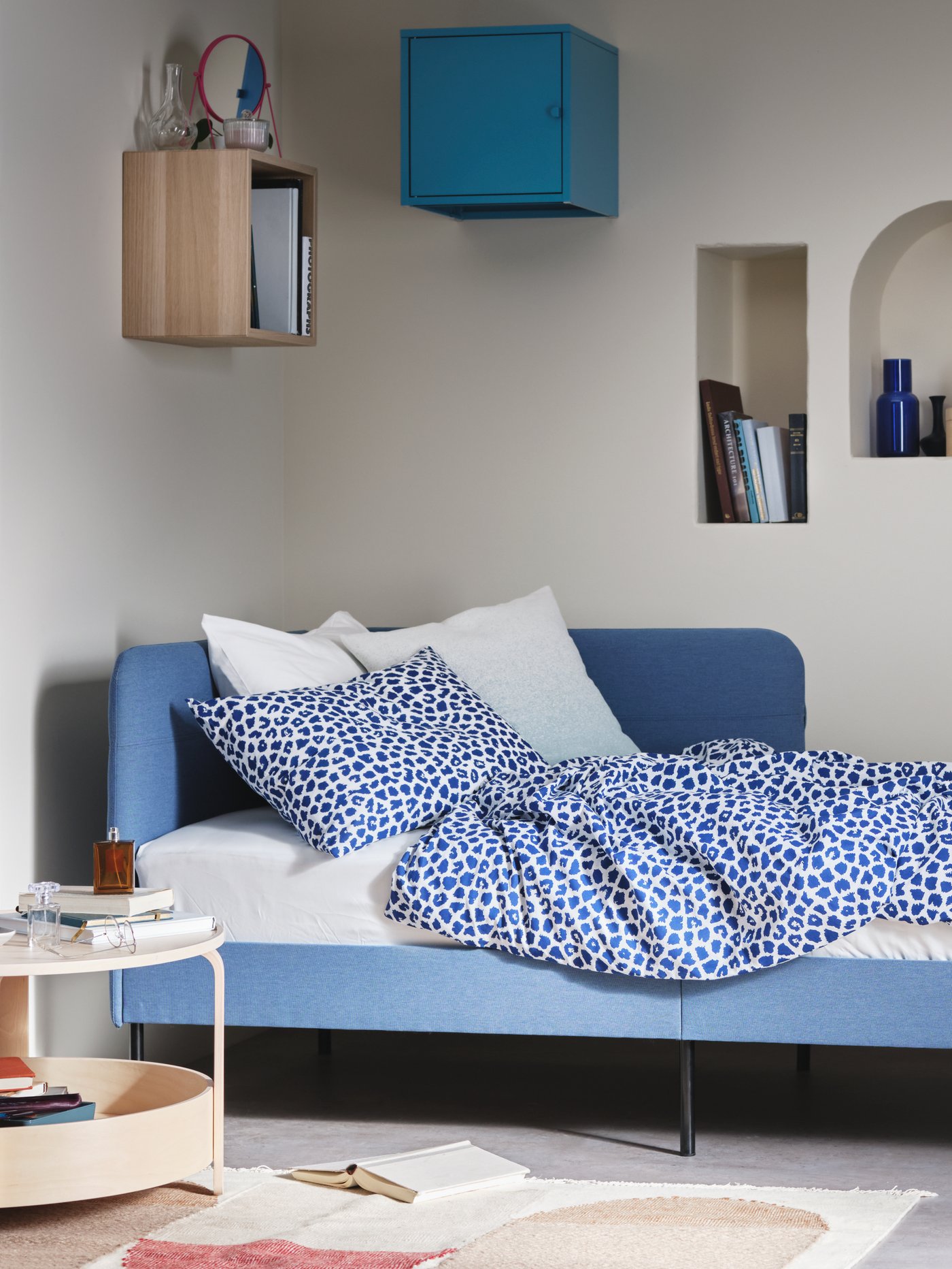 Dormitorio con estructura de cama tapizada con cabecero de esquina, juego de cama, mesa de centro, alfombra, armarios, libros, jarrón y espejo.