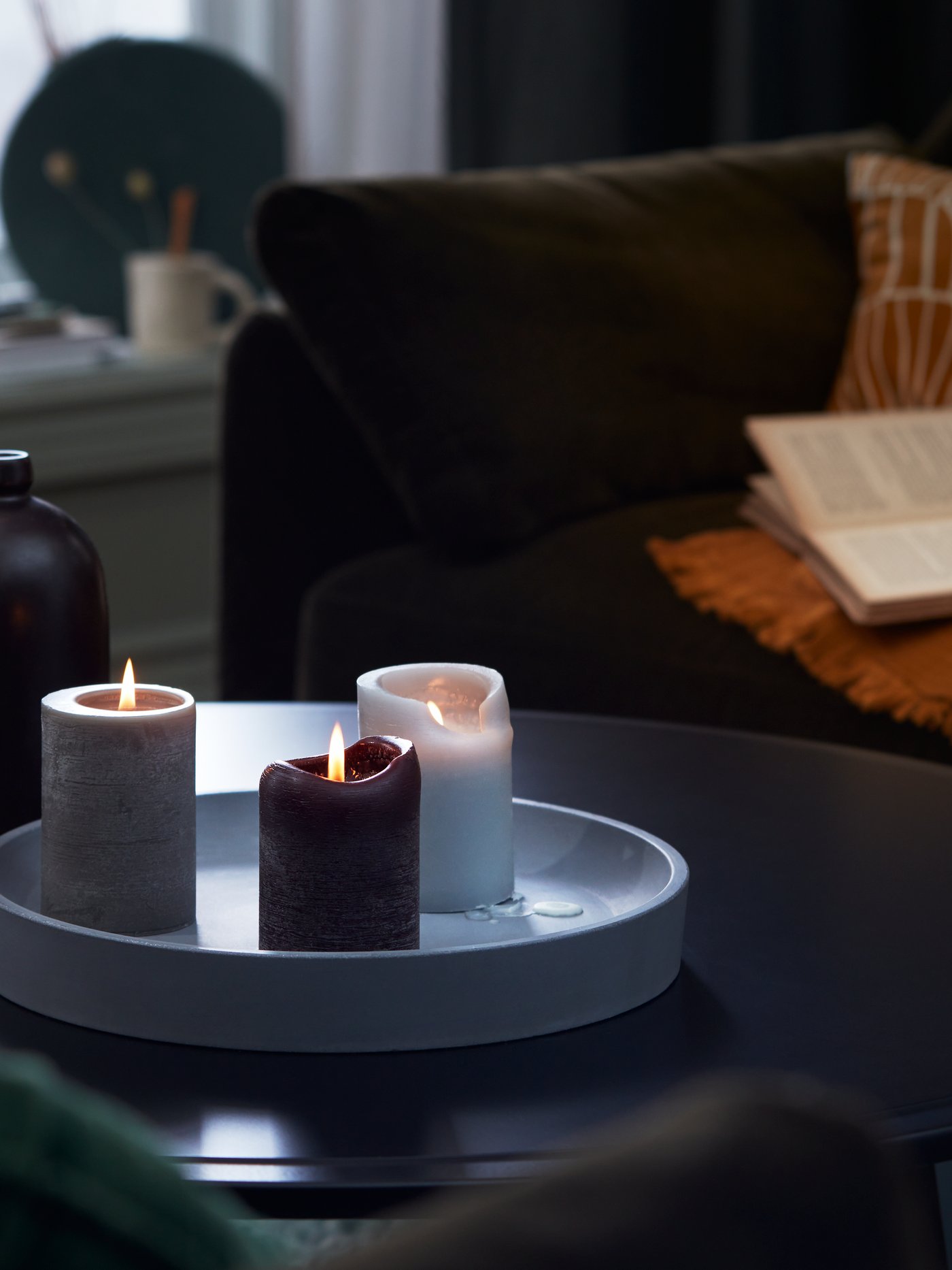Un plat à bougies BEGÄRAN gris foncé avec trois bougies parfumées BEHJÄRTAD allumées sur une table basse KRAGSTA dans un salon.