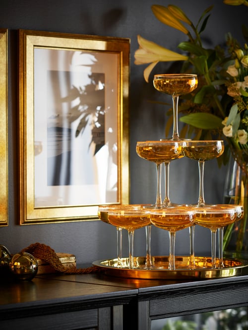 Une imposante pyramide festive en verre clair avec coupes à champagne STORHET et liquide doré se dresse sur un plateau cuivré au-dessus d’un meuble.