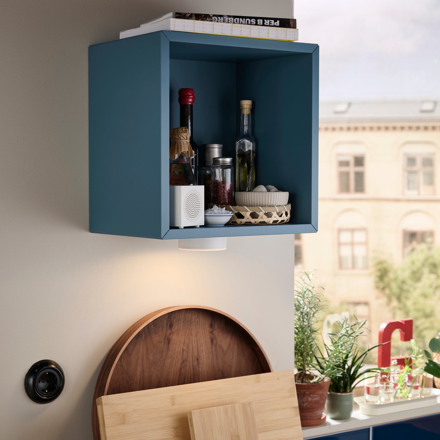 A dark grey-blue EKET wall-mounted shelving unit is placed on a kitchen wall and has bottles and jars stored inside it.