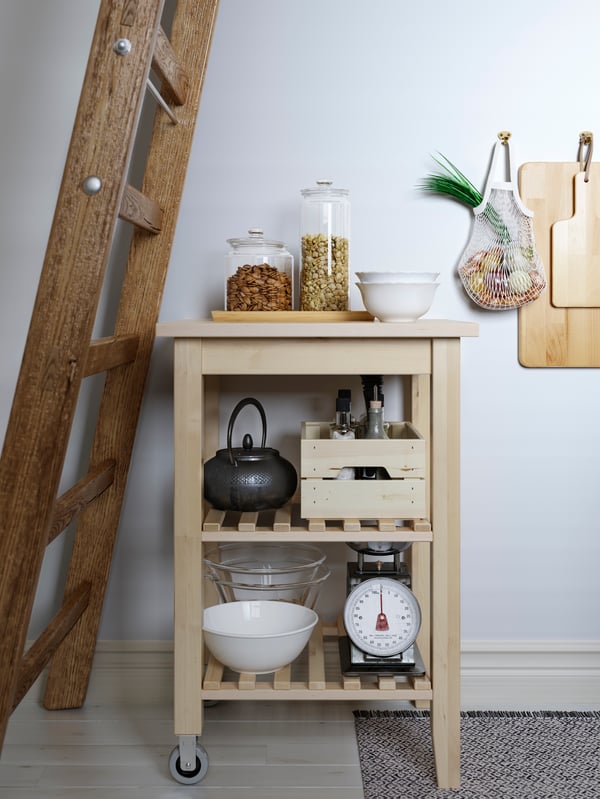 A kitchen trolley in birch stands under a wooden ladder. Kitchen utensils, jars and a teapot are on the trolley shelves.
