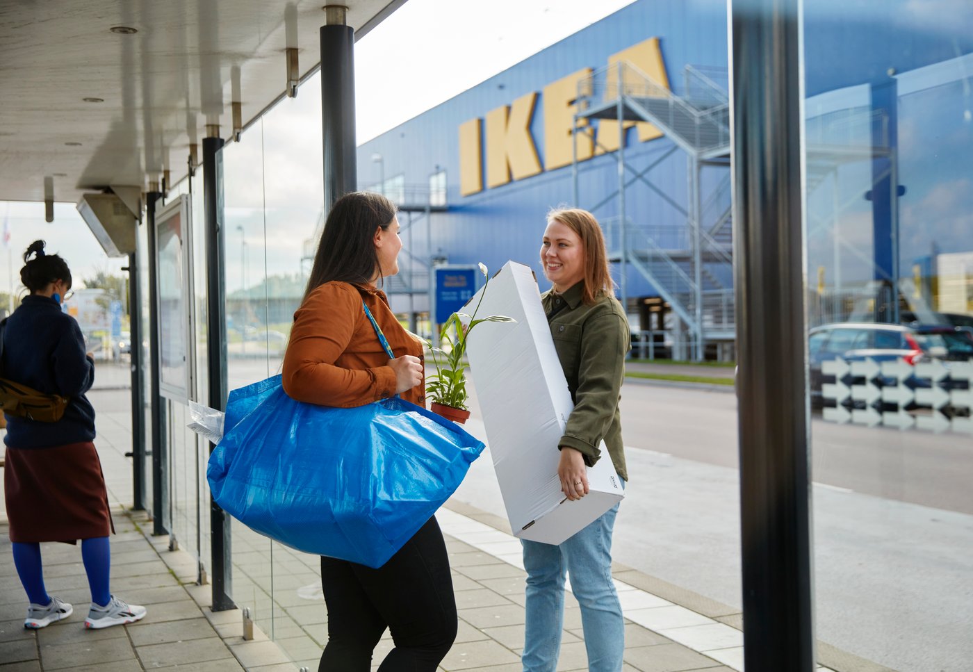 Kaksi naista jotka seisovat IKEA-tavaratalon ulkopuolella kantaen ostoksia