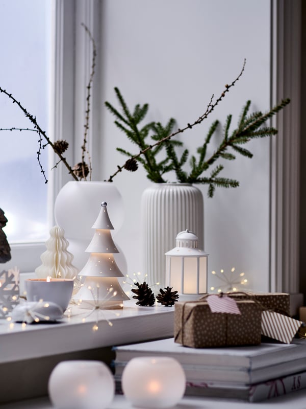 Fönster pyntat för novent med små granar, ljuslyktor och grenar i vas på ett bord vid ett fönster.