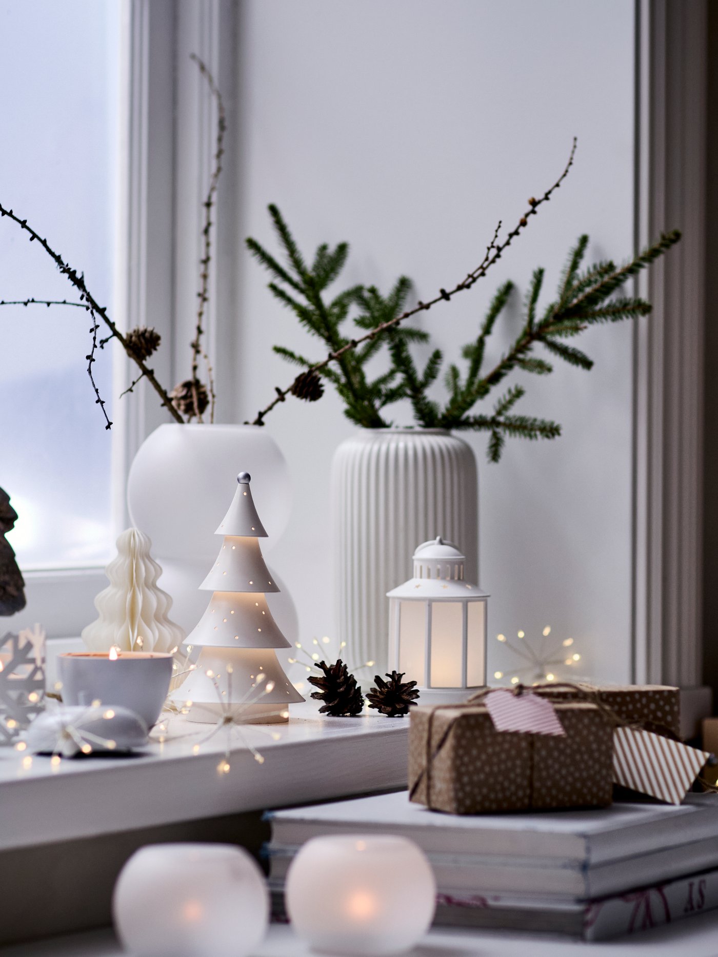 Fönster pyntat för novent med små granar, ljuslyktor och grenar i vas på ett bord vid ett fönster.
