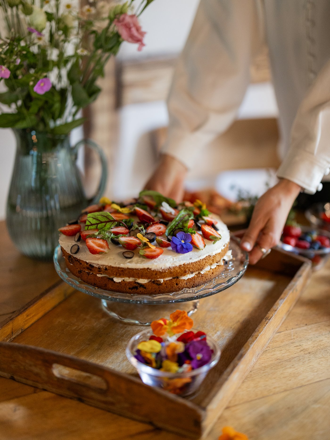 Eine Person präsentiert eine selbstgebackene Torte, die mit frischen Blumen auf einem Holztisch dekoriert ist, was eine einladende und rustikale Ästhetik schafft.