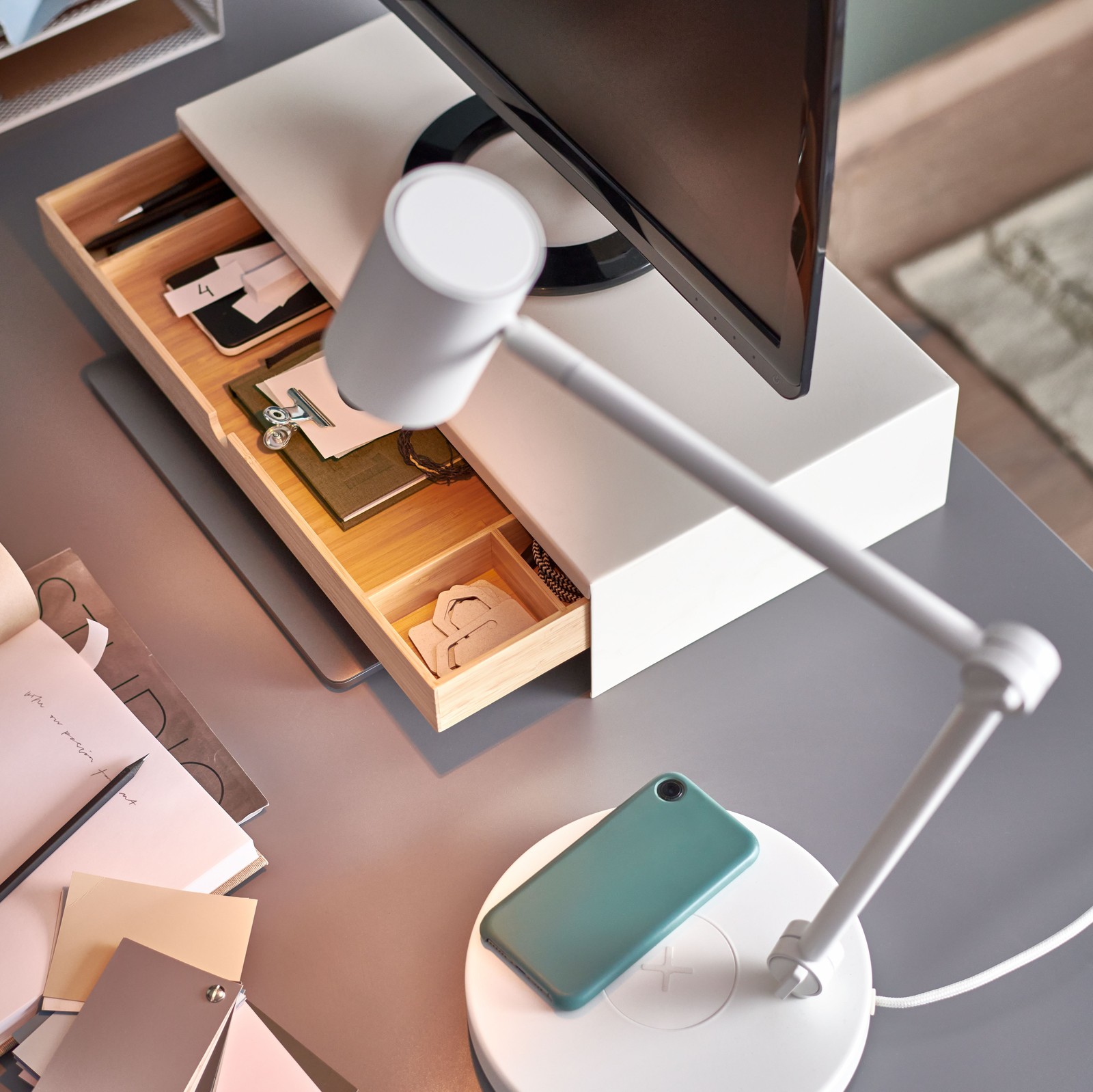 O veioză LED cu încărcare wireless NYMÅNE și monitorul unui computer pe un suport pentru monitor cu un sertar deschis.