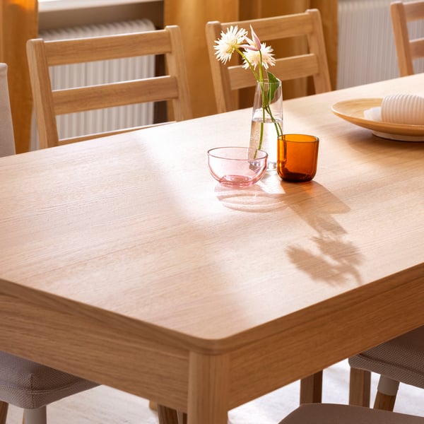 A light wood-toned TONSTAD table setting featuring a vase with flowers, glasses, and a plate on a sunlit surface.