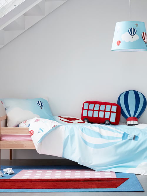 Child's bedroom with solid wood bed frame with guard rail, colourful rug, blue bed linens, blue lamp shade, and cushions.