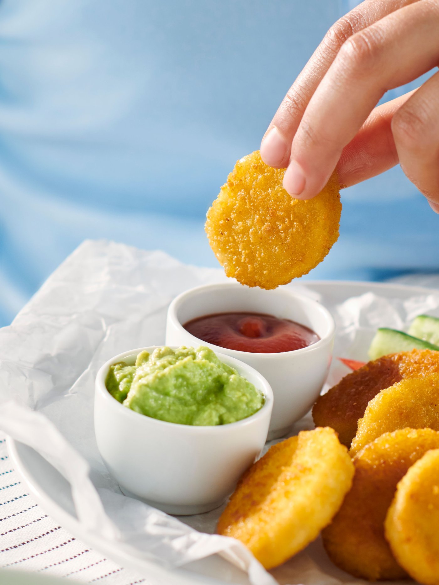 종이 위에 담긴 황금빛 감자 너겟을 손으로 집어 케첩 소스에 찍으려는 장면으로, 초록색 디핑 소스와 추가 감자 너겟이 함께 세팅된 이미지