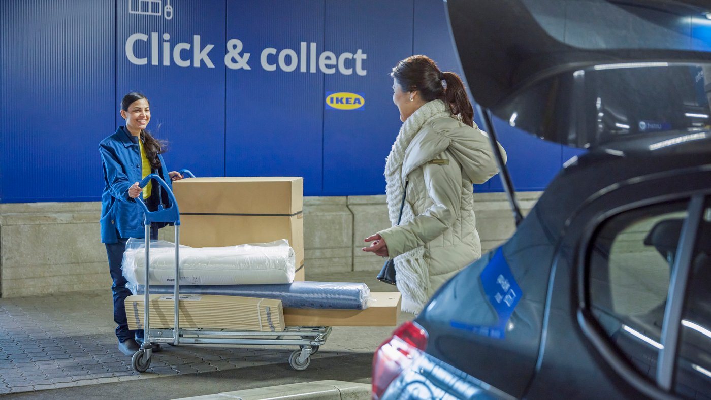 Una persona amb jaqueta blanca rep un carretó amb diversos paquets grans i llargs d'un empleat amb uniforme blau, a la zona de Click & Collect d'IKEA a la nit. Al fons es veu la paret blava amb el logotip d'IKEA i el text "Click & collect". A la dreta, hi ha obert el maleter d'un cotxe preparat per carregar els productes.