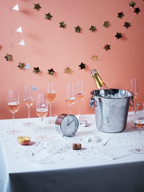 Des verres à champagne STORSINT sur une table recouverte de confettis avec une bouteille dans un seau à glace, une chaîne de décorations à étoile dorée et une horloge.