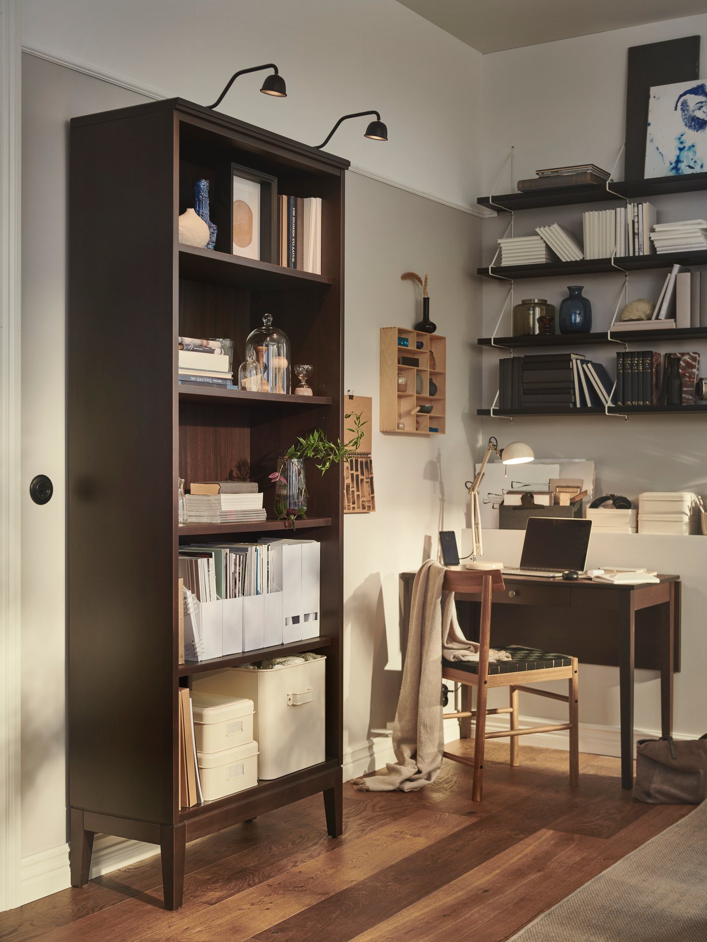Salón con una estantería IDANÄS marrón con cajas y revisteros, junto a una mesa abatible y una estantería expositora beige.