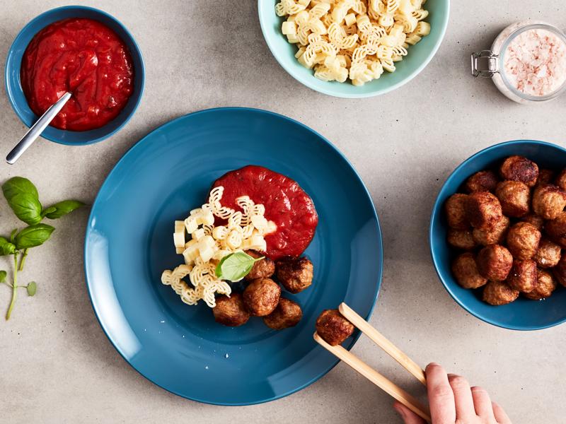Assiette bleu remplie de boulettes, pates, ketchup avec une main qui vient chercher une boulette avec une pince